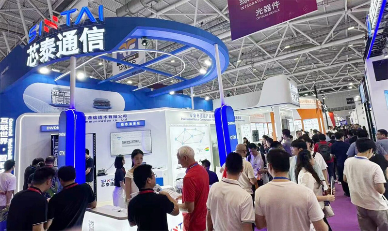  The SINTAI booth at a busy trade show. The brightly lit, blue and white booth is crowded with attendees viewing displays of optical communication products.